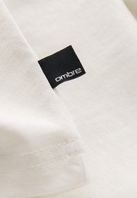 Tissu blanc avec une étiquette tissée noire comportant le texte « ombre » brodé en blanc, fixée près d’un bord ourlé.