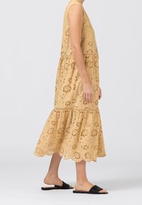 Un vestido sin mangas de color amarillo claro con patrones de ojales florales y un diseño en capas con un dobladillo con puntas. Llevado con sandalias negras de tipo deslice.