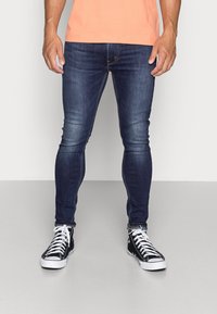 Hombre vistiendo jeans ajustados de color azul oscuro, zapatillas altas negras con cordones blancos y una camiseta naranja, de pie frente a un fondo liso.
