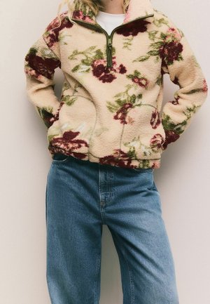 Person trägt einen cremefarbenen Fleecepullover mit roten und grünen Blumenmustern, einem Frontreißverschluss und blauen Jeans, die Hände in den Taschen.