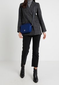 Blazer grigio a quadri con doppio petto indossato su un dolcevita grigio, abbinato a jeans neri tagliati e stivaletti neri. Borsa a tracolla blu con tracolla a catena.