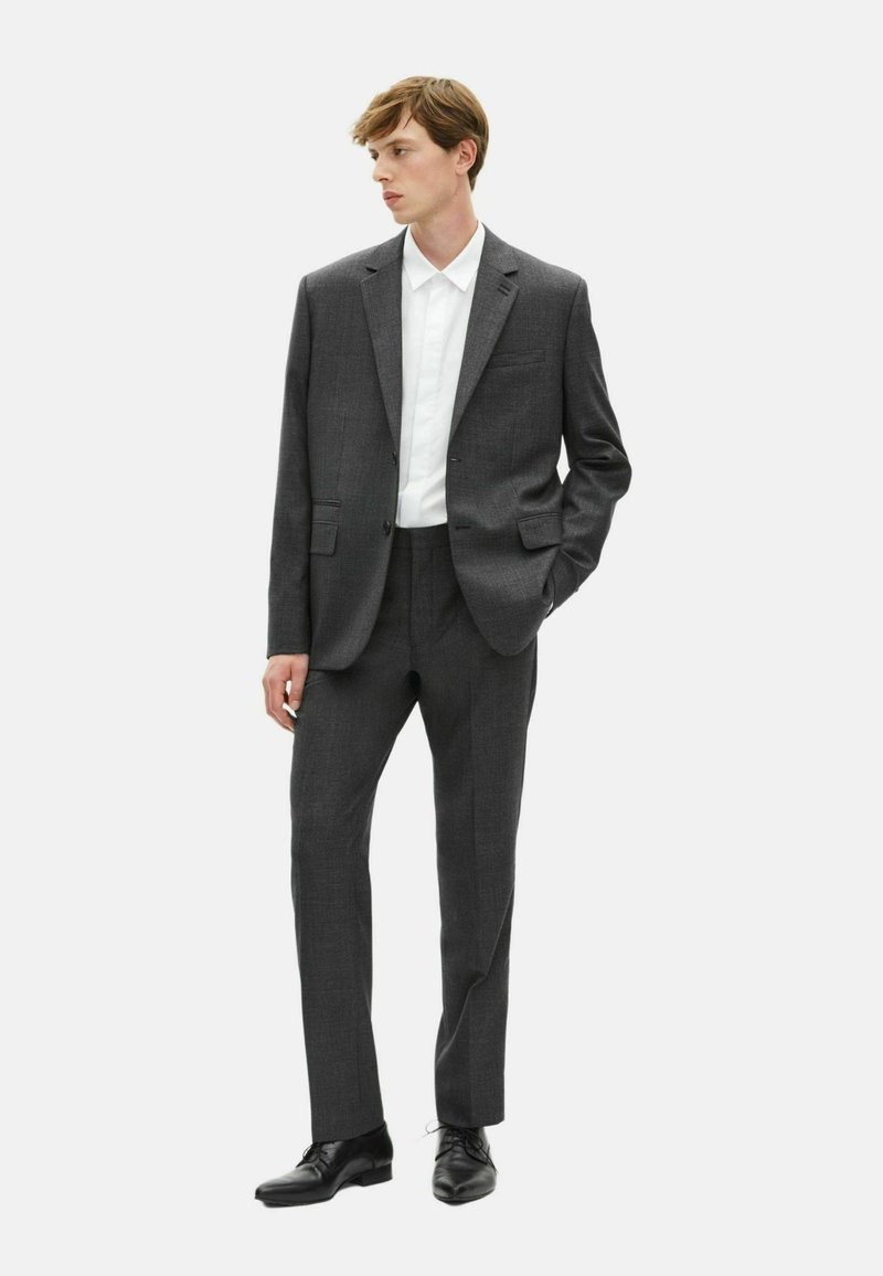 Grijze op maat gemaakte suit van gestructureerde stof, met een voorzak op de blazer, gedragen met een wit overhemd met kraag en zwarte schoenen.