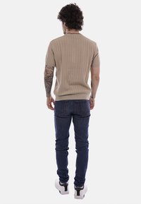 Uomo con capelli ricci e tatuaggi sulle braccia che indossa un maglione beige a maniche corte, jeans blu scuro e scarpe da ginnastica bianche, in piedi di spalle.