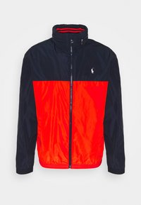 Väriestetty takki, jossa on tummansininen yläosa ja kirkkaan punainen alaosa, valmistettu sileästä, kevyestä kankaasta ja varustettu vetoketjulla.