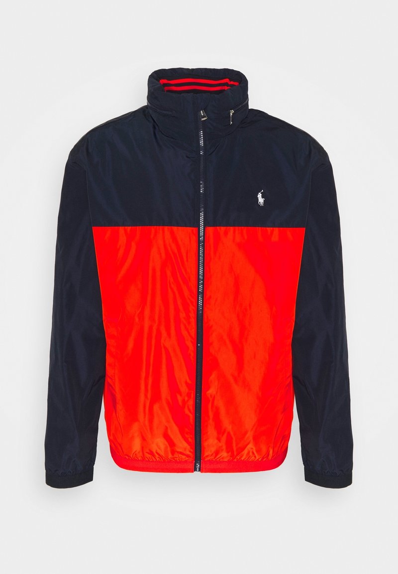 Väriestetty takki, jossa on tummansininen yläosa ja kirkkaan punainen alaosa, valmistettu sileästä, kevyestä kankaasta ja varustettu vetoketjulla.