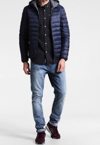 Marinblå puffjacka med huva, lätt design, grå inredning, kombinerad med en svart rutig skjorta, ljusblå jeans och burgundyröda sneakers.