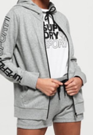 Personne portant un sweat à capuche zippé gris clair avec le texte "Superdry" sur les manches, un short assorti et un T-shirt blanc graphique.