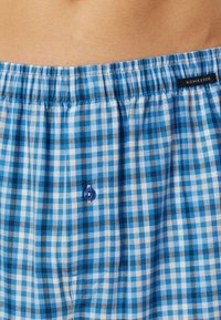 Pantaloni boxer a scacchi blu con elastico in vita, dotati di un piccolo bottone blu nella parte anteriore e una texture di tessuto morbido.