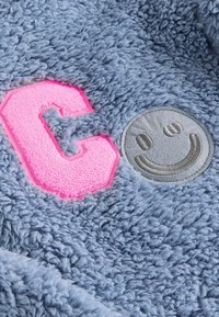 Puffet blå stof har et pink broderet bogstav "C" og et gråt smilende ansigtspatch med syede detaljer. Blød tekstur og legende design.