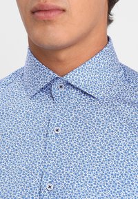 Niebieska koszula zapinana na guziki z wzorem drobnych białych kwiatów, kontrastowymi guzikami i klasycznym kołnierzem o gładkiej fakturze tkaniny.