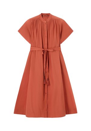 Femme portant une robe ceinturée orange rouille, tenant un sac tissé noir et crème, chaussée de talons ouverts, debout devant un fond beige uni.