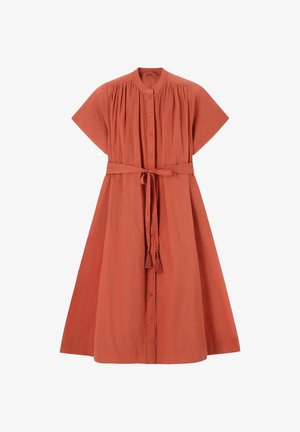 Femme portant une robe ceinturée orange rouille, tenant un sac tissé noir et crème, chaussée de talons ouverts, debout devant un fond beige uni.