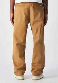 Beżowe bawełniane spodnie o prostym kroju, z dwoma tylnymi kieszeniami i widoczną metką. Noszone z białymi trampkami.