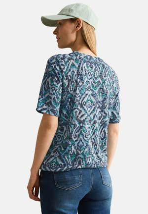 Kortærmet skjorte med et blåt og grønt abstrakt mønster, lavet af letvægtstof. Sammen med en lysegrøn kasket og denim jeans.