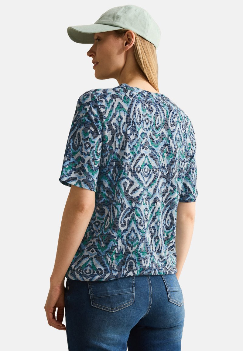 Maglietta a maniche corte con un motivo astratto blu e verde, realizzata in tessuto leggero. Abbinata a un cappellino verde chiaro e jeans in denim.