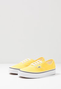 Žluté plátěné tenisky s bílými tkaničkami a gumovou podrážkou. Vyznačují se černým pruhem kolem spodního okraje a štítkem s logem na boku.