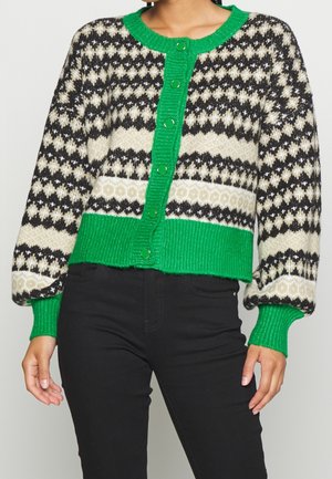 Cardigan en tricot à motif noir et blanc avec patte de boutonnage, col, poignets et ourlet verts vifs, porté avec un pantalon noir.