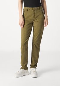 Olive spalvos liesi kostiumai, pagaminti iš minkštos medžiagos, su užtrauktukais kišenėmis ir lygia textura. Dėvimi su baltais sportbačiais.