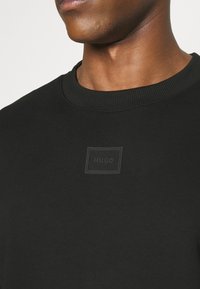 Gros plan d'une personne portant un sweat-shirt noir à col rond avec un logo carré embossé subtil marqué "HUGO" sur la poitrine.