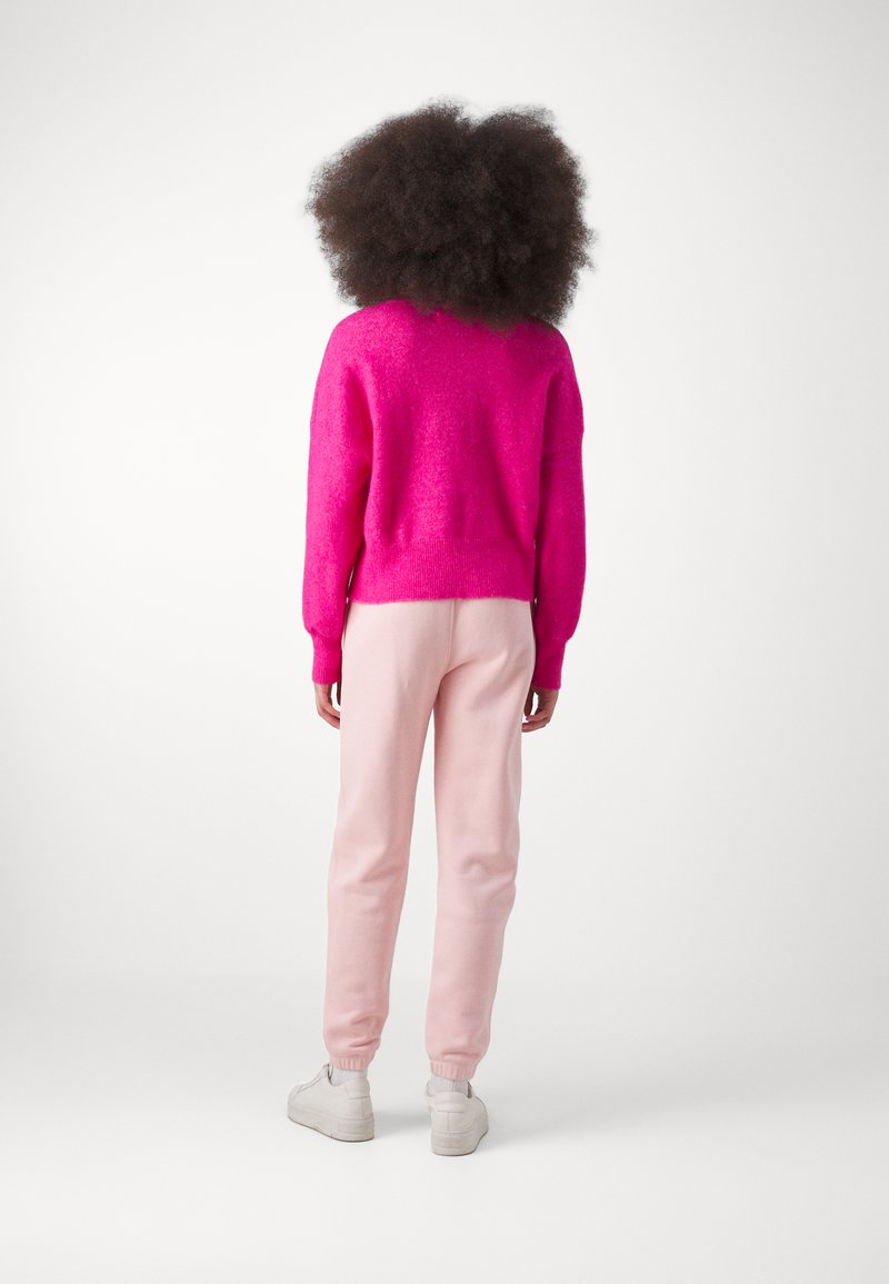 Un pull en peluche rose, un pantalon de survêtement rose clair et des baskets blanches. Le pull a une coupe décontractée et des poignets côtelés, avec une texture douce.