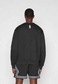 Homem de costas, vestindo um moletom da NBA preto e calções de basquete pretos com detalhes brancos, contra um fundo claro e liso.