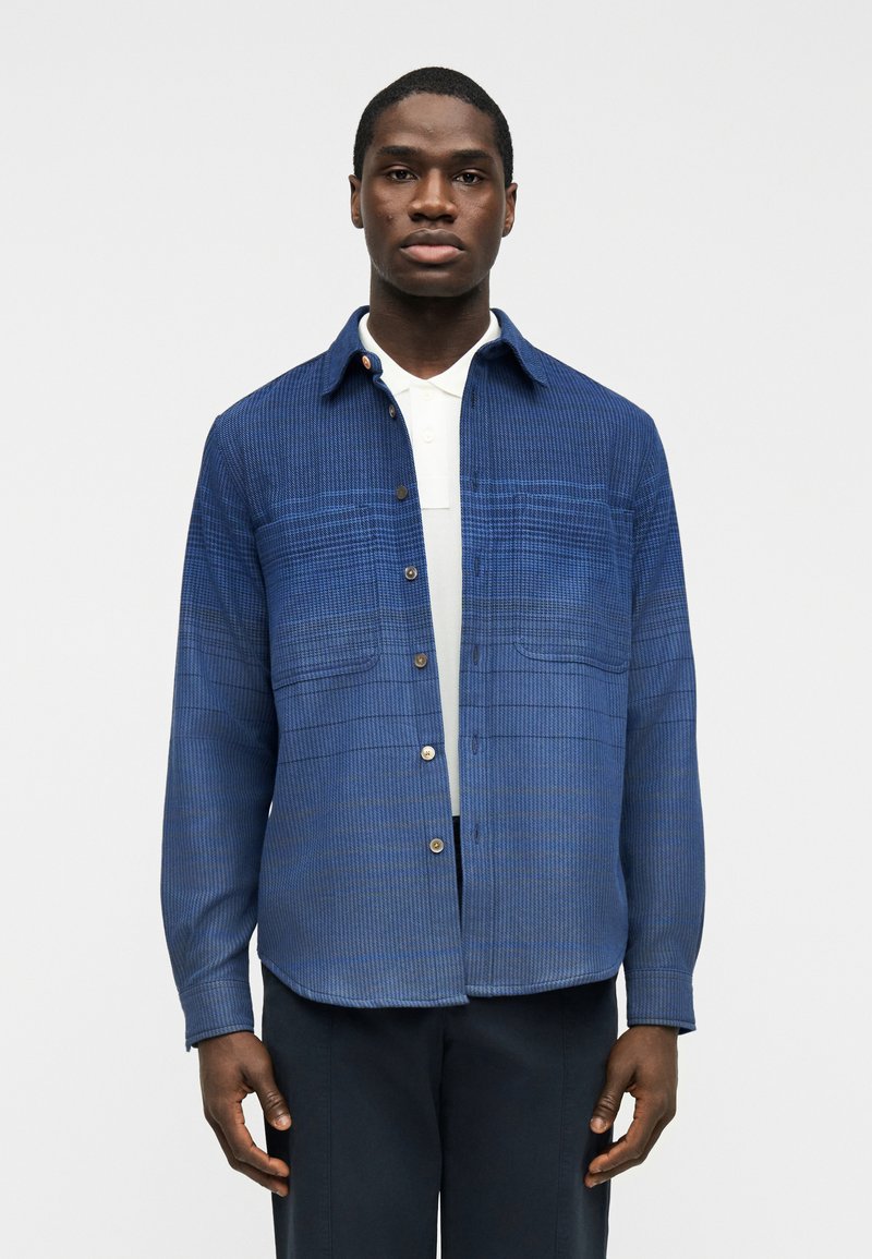 Άνδρας με μπλε πουκαμισό με καρό κουμπιά πάνω από λευκό πόλο, στέκεται μπροστά σε απλό φωτεινό φόντο, κοιτώντας ευθεία.