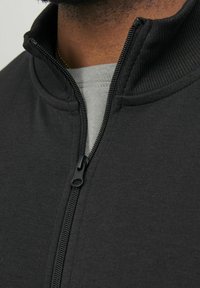 Chaqueta negra con cremallera y cuello alto, que presenta una textura de tela suave y una cremallera resistente. Superpuesta sobre una prenda de color gris claro.