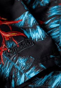 Tessuto nero con un motivo di foglie tropicali blu e rosse; include un logo "Superdry" ricamato in nero. La texture appare liscia.
