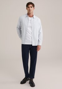 Šviesiai mėlynas marškinių tipo švarkas su subtiliu raštu, dėvimas ant balto marškinėlio, derinamas su tamsiai mėlynomis kelnėmis ir juodais raišteliais.