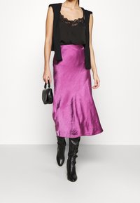 Femme portant un haut noir orné de dentelle, un pull sans manches noir sur les épaules, une jupe midi violette brillante, des bottes noires et tenant un petit sac à main noir.