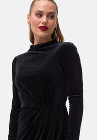 Czarna sukienka z weluru z długimi rękawami, wysokim dekoltem i drapowaniem w talii. Lśniące dodatki to kolczyki w kształcie kropli.