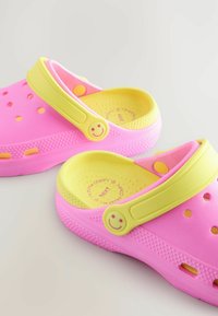 Roze plastic klompen met een gele band, een smiley-knopaccent en ventilatiegaatjes. Gestructureerd oppervlak voor grip en comfort.