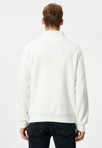 Man met kort bruin haar, gekleed in een eenvoudige witte langbeenpullover en donkere jeans, staande met de rug naar de camera tegen een witte achtergrond.