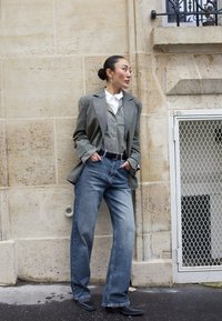 Pilkos vilnos švarkas per dryžuotą liemenę ir baltą marškinius, derinamas su džinsais ir juodais kulkšninių botais. Stovi prieš akmeninę sieną.