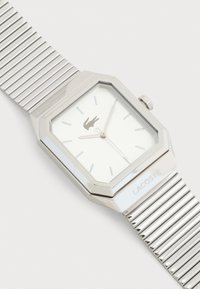 Ασημένιο ορθογώνιο ρολόι χειρός με μεταλλικό λουρί, μινιμαλιστικό καντράν, ασημένιες ενδείξεις ώρας και δείκτες, και λογότυπο Lacoste στο καντράν και το λουρί.