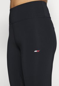 Czarne legginsy sportowe z wysokim stanem, z małym logo w kolorach czerwonym, białym i niebieskim po boku oraz teksturowymi akcentami przy szwach.