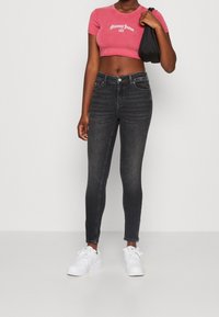 Mujer vistiendo una camiseta recortada rosa de Tommy Jeans, jeans ajustados negros, zapatillas blancas y sosteniendo un bolso de hombro negro.