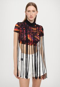 Top corto con un estampado vibrante y multicolor en naranja, púrpura y negro, acentuado por largos flecos negros. Combinado con pantalones cortos blancos.
