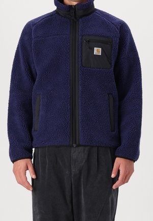 Veste polaire bleu marine avec fermeture éclair noire, poche poitrine noire avec fermeture éclair et écusson logo, portée avec un pantalon en velours côtelé gris foncé.