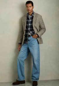 Grauer strukturierter Blazer über einem dunklen karierten Hemd, kombiniert mit hellblauen, lockeren Jeans und dunkelbraunen Schuhen. Das Model steht vor einem neutralen Hintergrund.