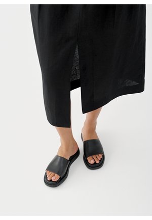 Stopy w czarnych sandałach typu slip-on z odkrytymi palcami i grubymi paskami, zestawione z długą czarną spódnicą z rozcięciem z przodu na białym tle.