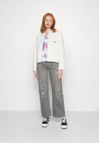 Vit jeansjacka, blommig pastellskjorta, grå cargobyxor med färgsplatter, och svarta sneakers med vita detaljer.