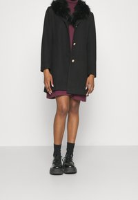 Manteau noir à col en fausse fourrure, avec deux boutons, coupe décontractée, porté sur une robe bordeaux, associé à des chaussures et des chaussettes noires.