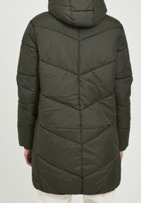 Osoba v tmavozeleném prošívaném zimním kabátu s kapucí, viděná zezadu, s viditelnými světlými kalhotami.