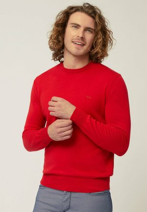 Maglione rosso lavorato a maglia con scollatura a girocollo, polsini e orlo a costine, caratterizzato da un piccolo logo sul petto, indossato sopra pantaloni grigio chiaro.