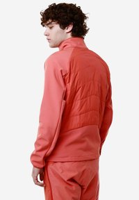 Giacca sportiva corallo con colletto alto e pannelli testurizzati cuciti sul retro. Maniche lunghe e design aderente nello stesso colore.