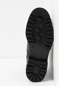 Suela de goma negra texturizada con surcos profundos y un número de modelo 37. La parte superior cuenta con un panel lateral con detalle de patrón en relieve.