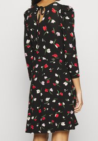 Czarna sukienka w czerwony, biały i różowy kwiatowy wzór, z długimi rękawami, z dekoltem w kształcie łezki i wiązaniem, noszona przez osobę pokazaną z tyłu.