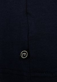 Mørkeblå bomuldsstof med ribbet tekstur, med et lille sort mærke med et hvidt "m" logo syet ved sømmen.