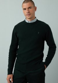 Dunkelgrüner Strickpullover mit strukturiertem Rippmuster, Rundhalsausschnitt und langen Ärmeln. Mit einem dezenten Logo auf der Brust und enganliegenden Bündchen.